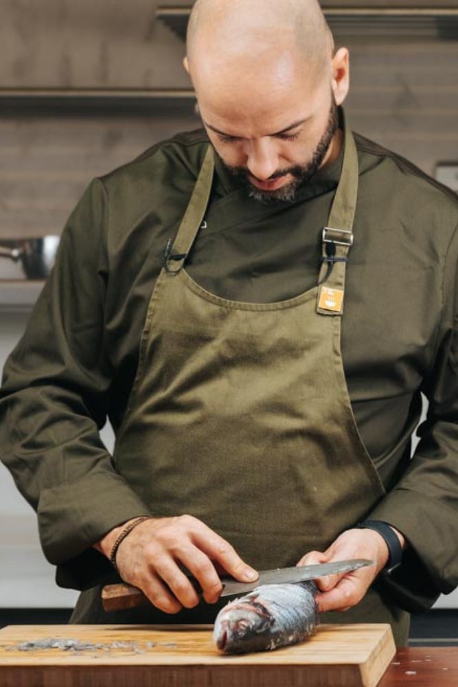 Justin Horne working at the pass — green apron, knife in hand, breaking down a whole fish for his No Waste Fish practice.